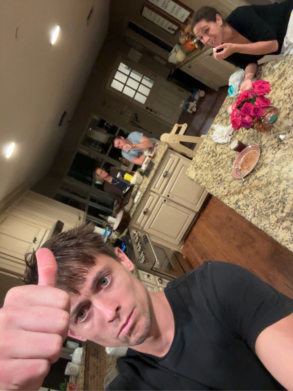 A playful kitchen selfie with a thumbs-up in the foreground and family in the background.